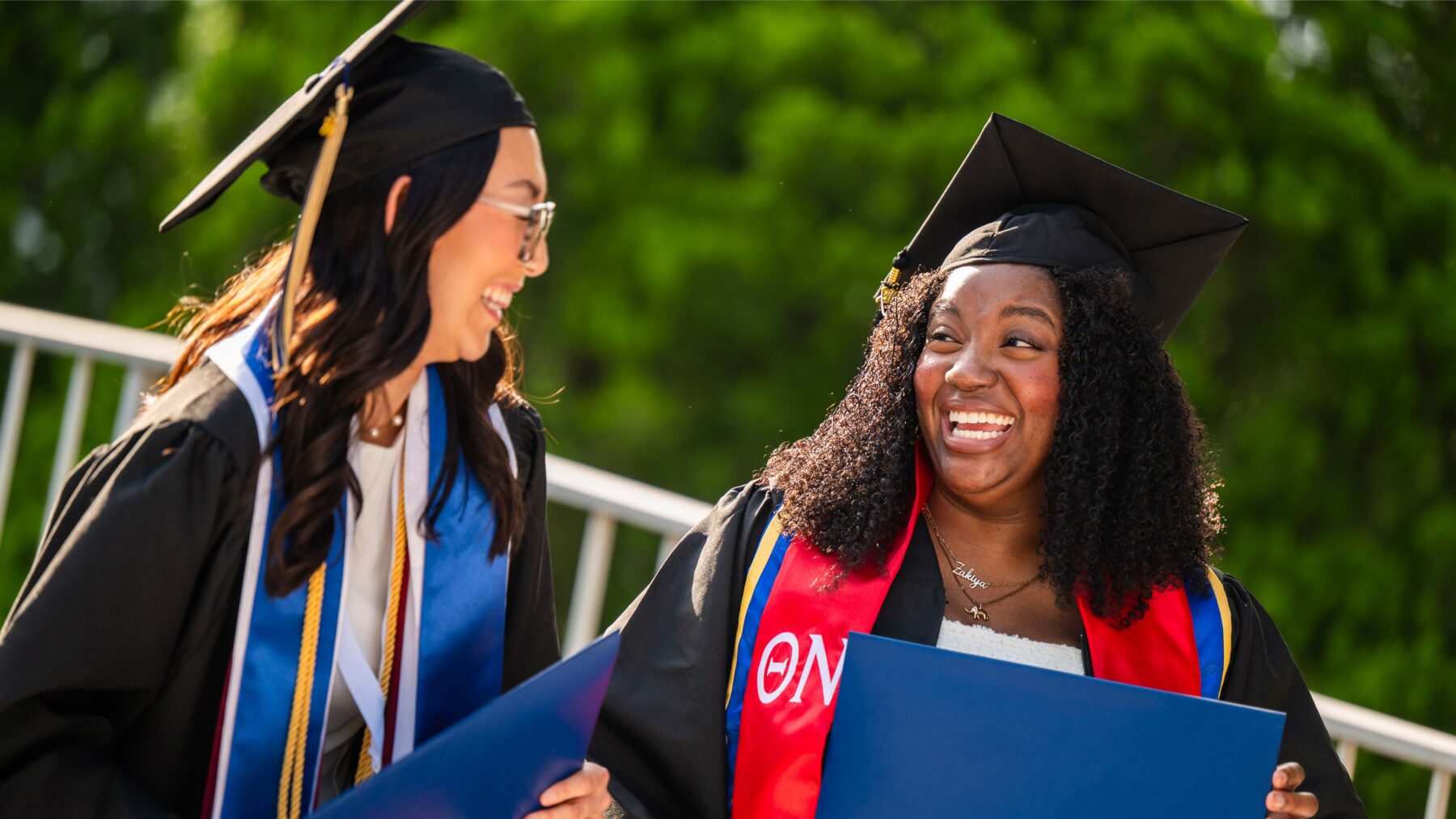 Two graduates smile at each other. Both are wearing graduation gowns and cords. The one on the right is holding a blue diploma cover.