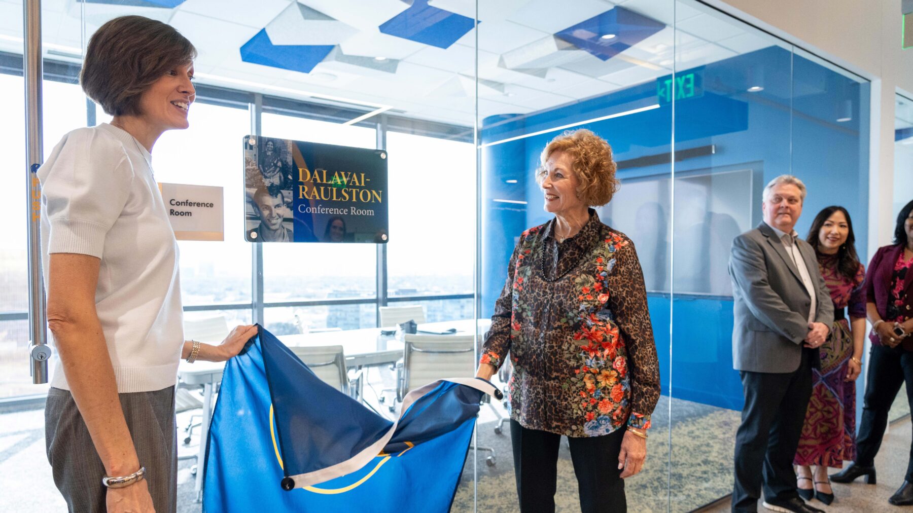 Julie Dalavai and a member of her family pull a blue flag off of a sign on a glass wall. The sign reads "Dalavai-Raulston Conference Room." Behind the glass wall, a conference room with a table and chairs is shown against a glass-walled backdrop showing panoramic views of the city.