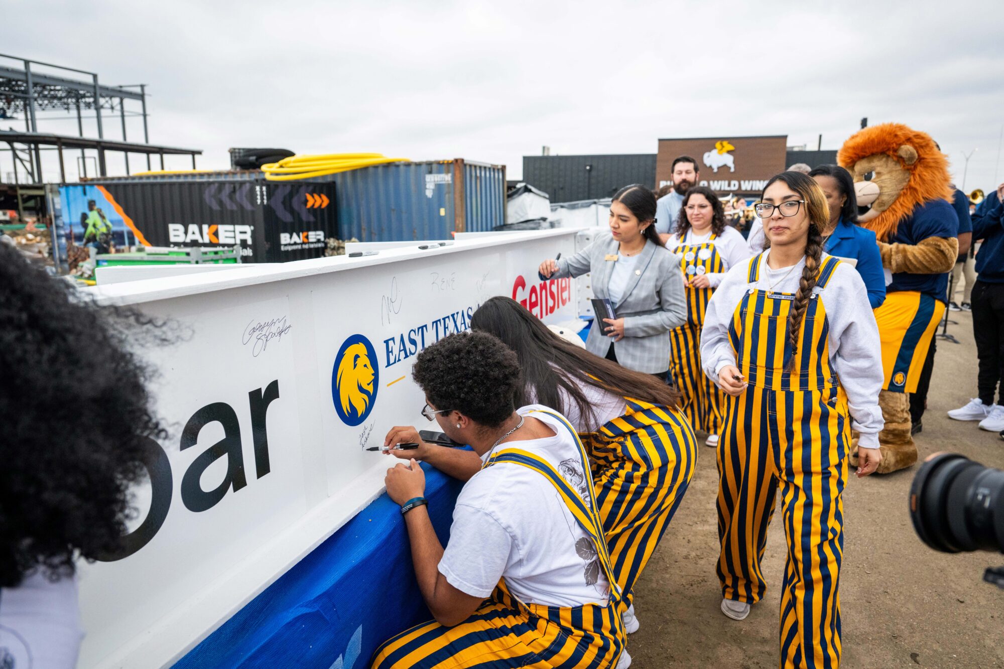 Students in gold and blue striped overalls sign their names on a large beam.