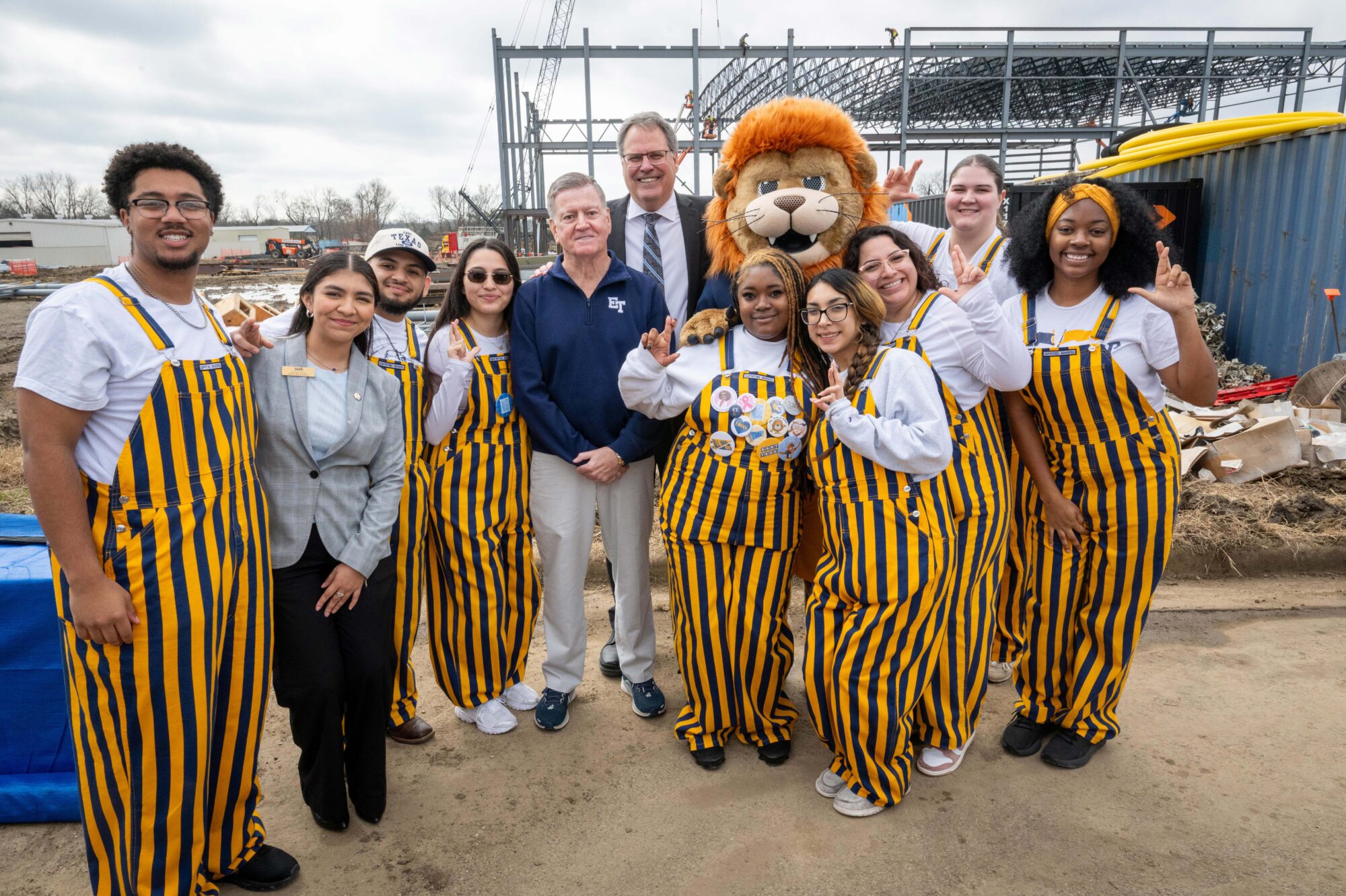 Eleven students stand with Lucky the Lion (the school mascot) in front of the construction project. They smile for the camera and make an "L" shape with their fingers.