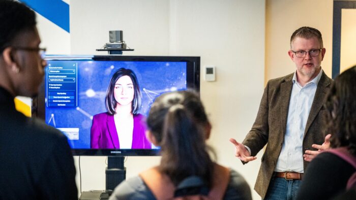 A person speaking to a crowd of people near a screen displaying a digital AI assistant.