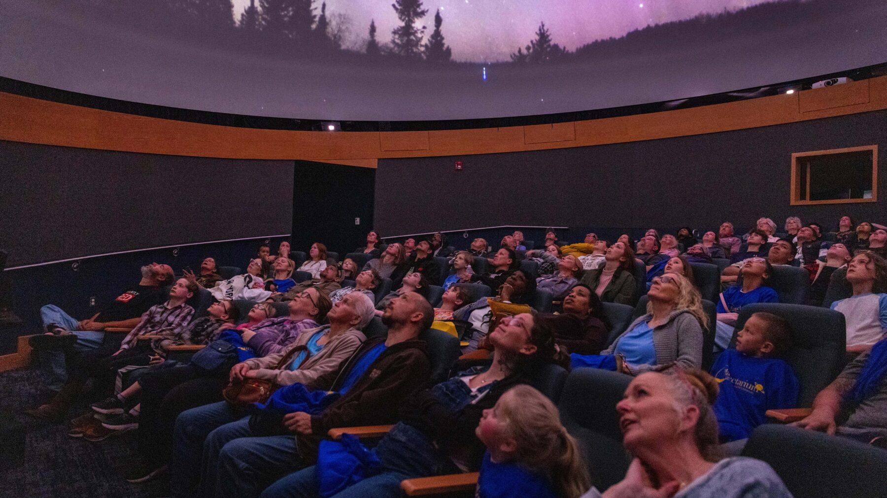 A crowd of people sitting in the Planetarium, watching the domed screen.