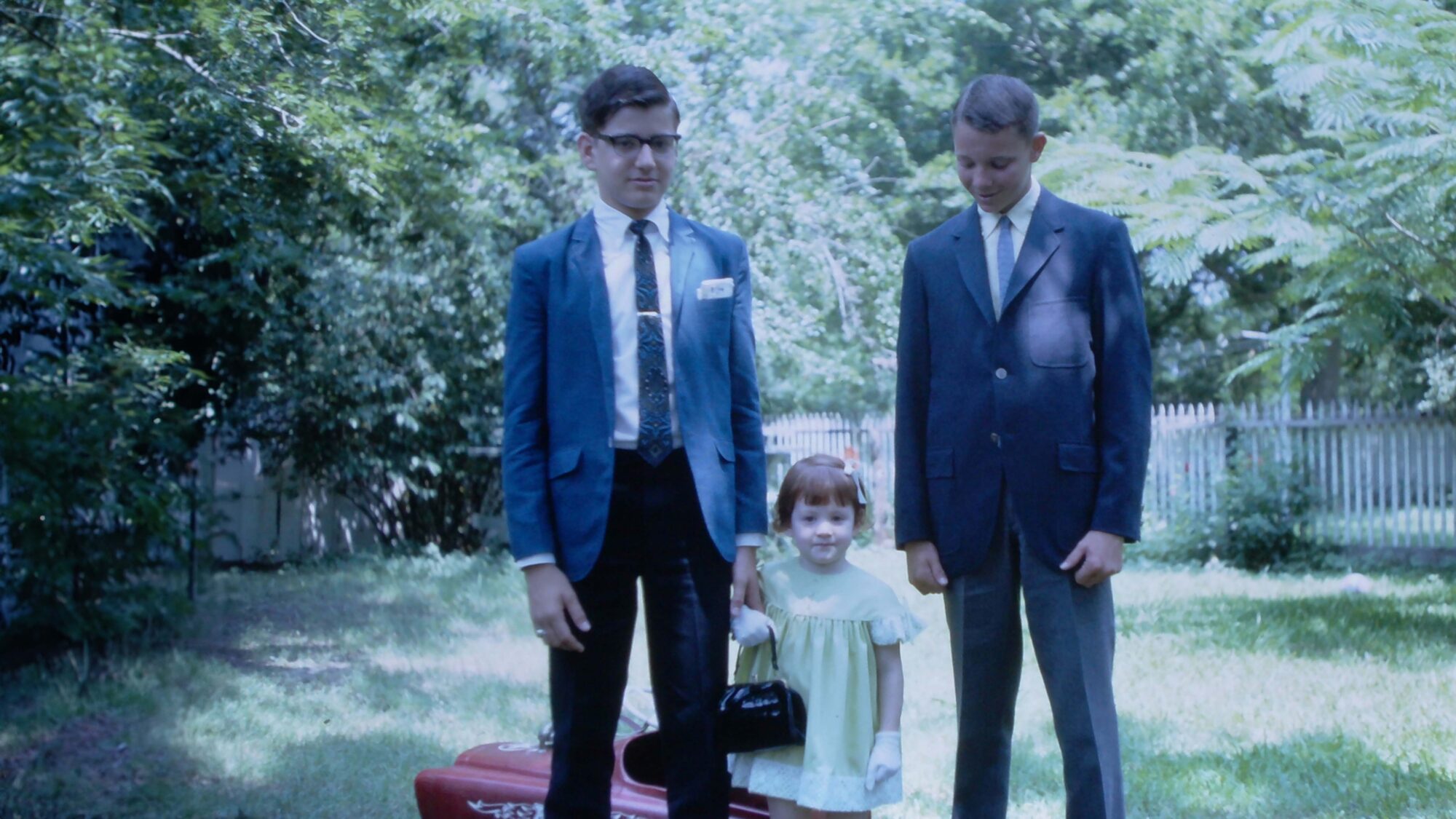 Three children stand in a yard with a white picket fence behind them. Two of the children are wearing suit jackets and the smaller child in the middle is wearing a light green dress while holding a black purse in a white-gloved hand.
