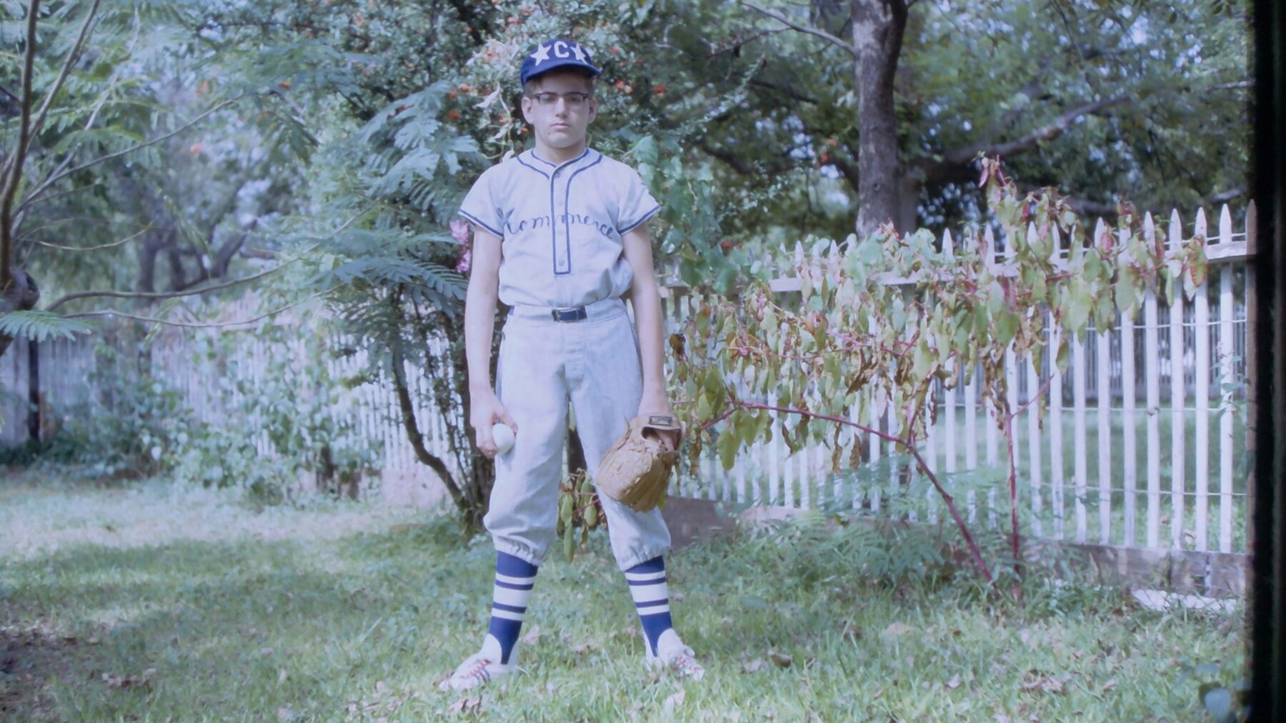 A child in a baseball uniform faces the camera. He has a baseball glove in one hand and a ball in the other.