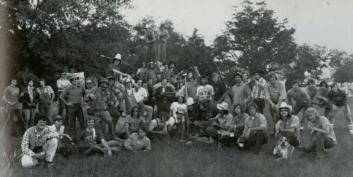 A black and white photo shows a group of young college men out in a field. Many are holding sticks and wearing cowboy hats. There are trees in the background.
