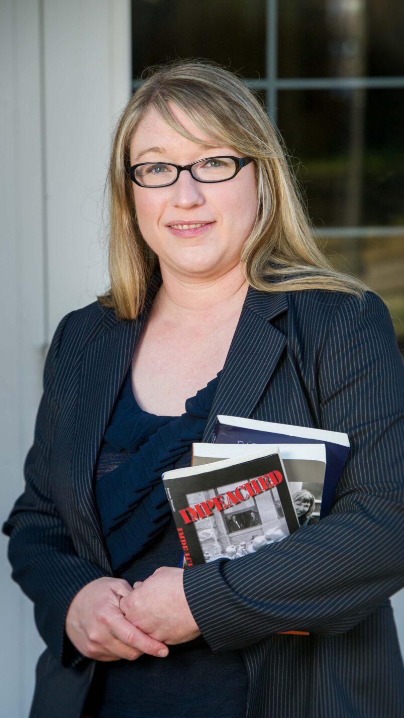 Dr. Jessica Brannon-Wranosky smiles at the camera while holding three books in her arm. Her hands are clasped together and she's wearing a black suit jacket.