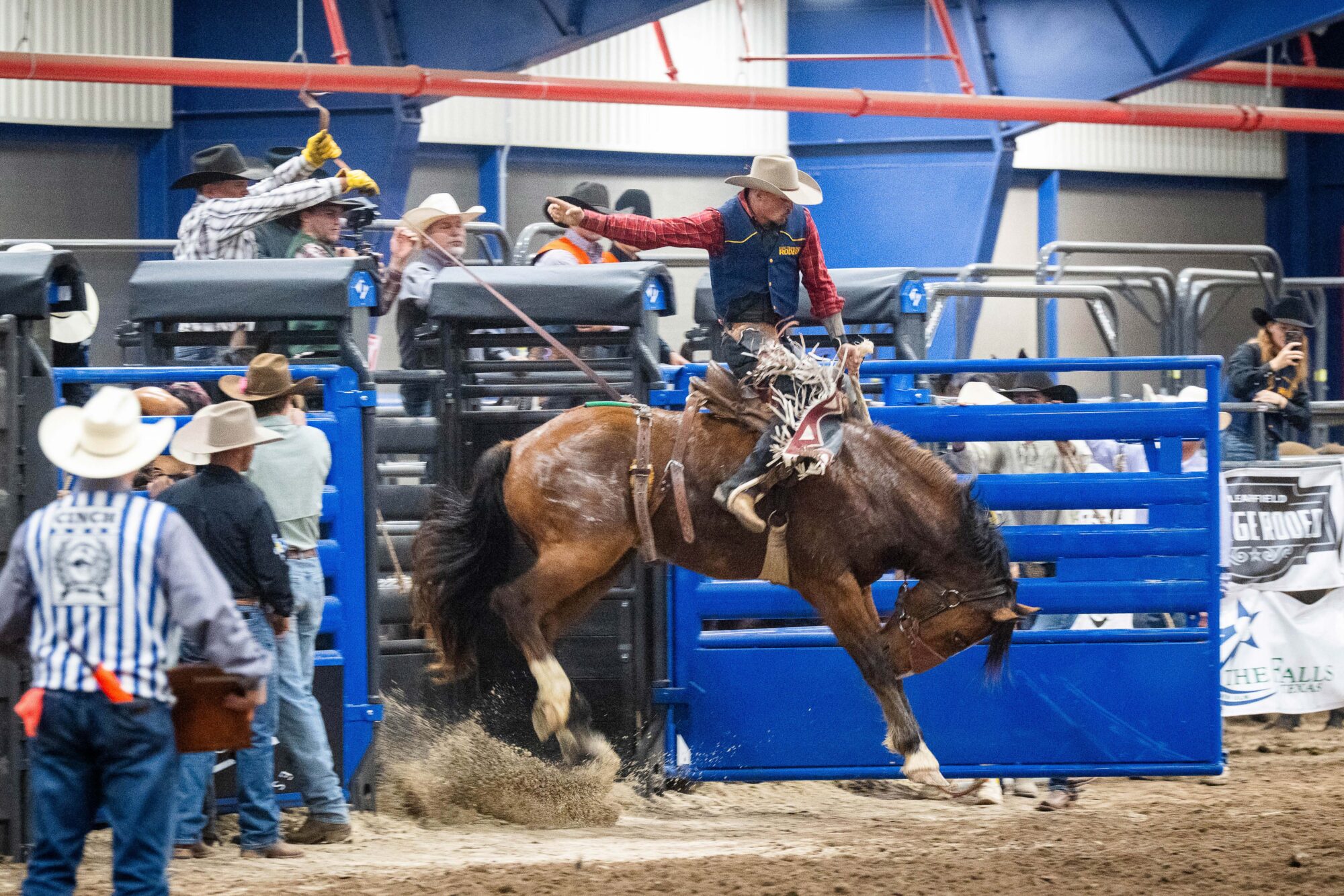 A cowboy rides a bucking horse. The cowboy is holding on with his left arm and holding his right arm out to his side. The horse's head is down and his back legs are kicking up.