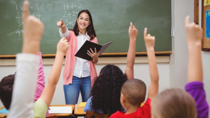 A female teacher standing at the head of a class while young students raise their hands.