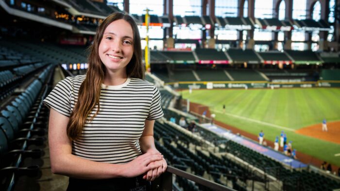 Ruby Blaylock smiles at the camera with the inside of the Texas Rangers stadium as the backdrop. The photo shows the field on the right and seating on the left of the photo.