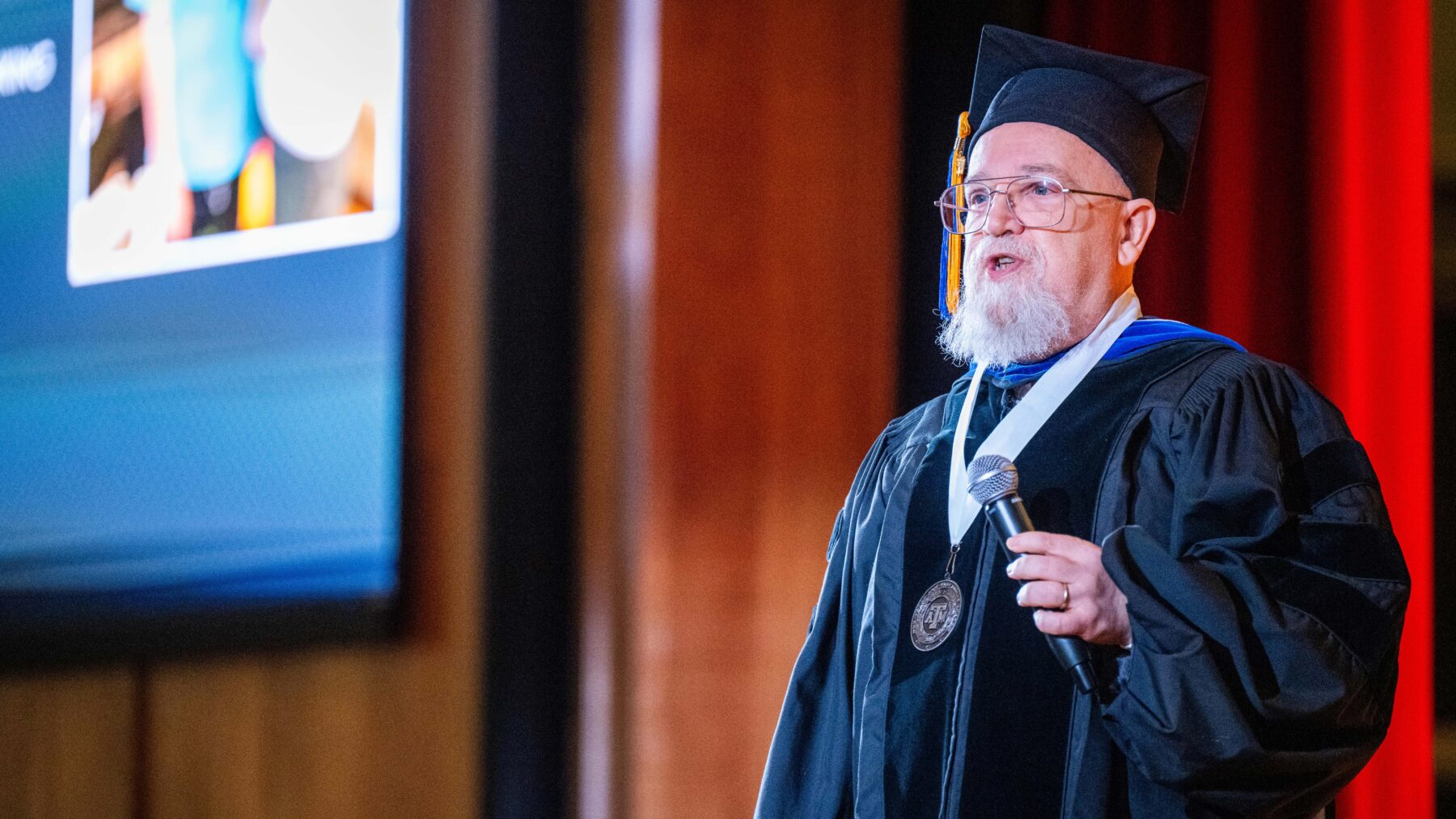 Man standing in academic regalia, with microphone in his hand.