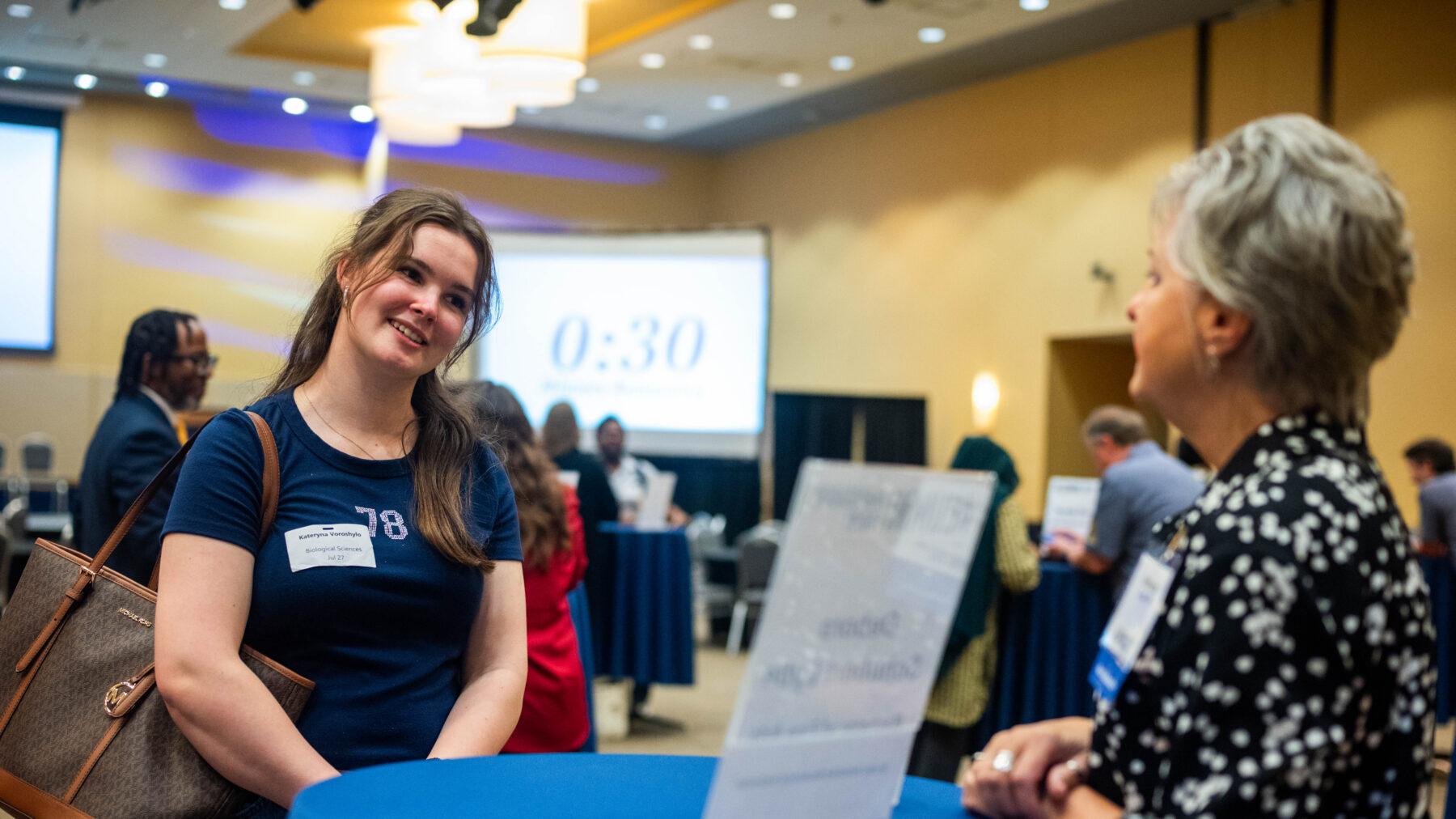 A student smiling in front of a woman talking to her.