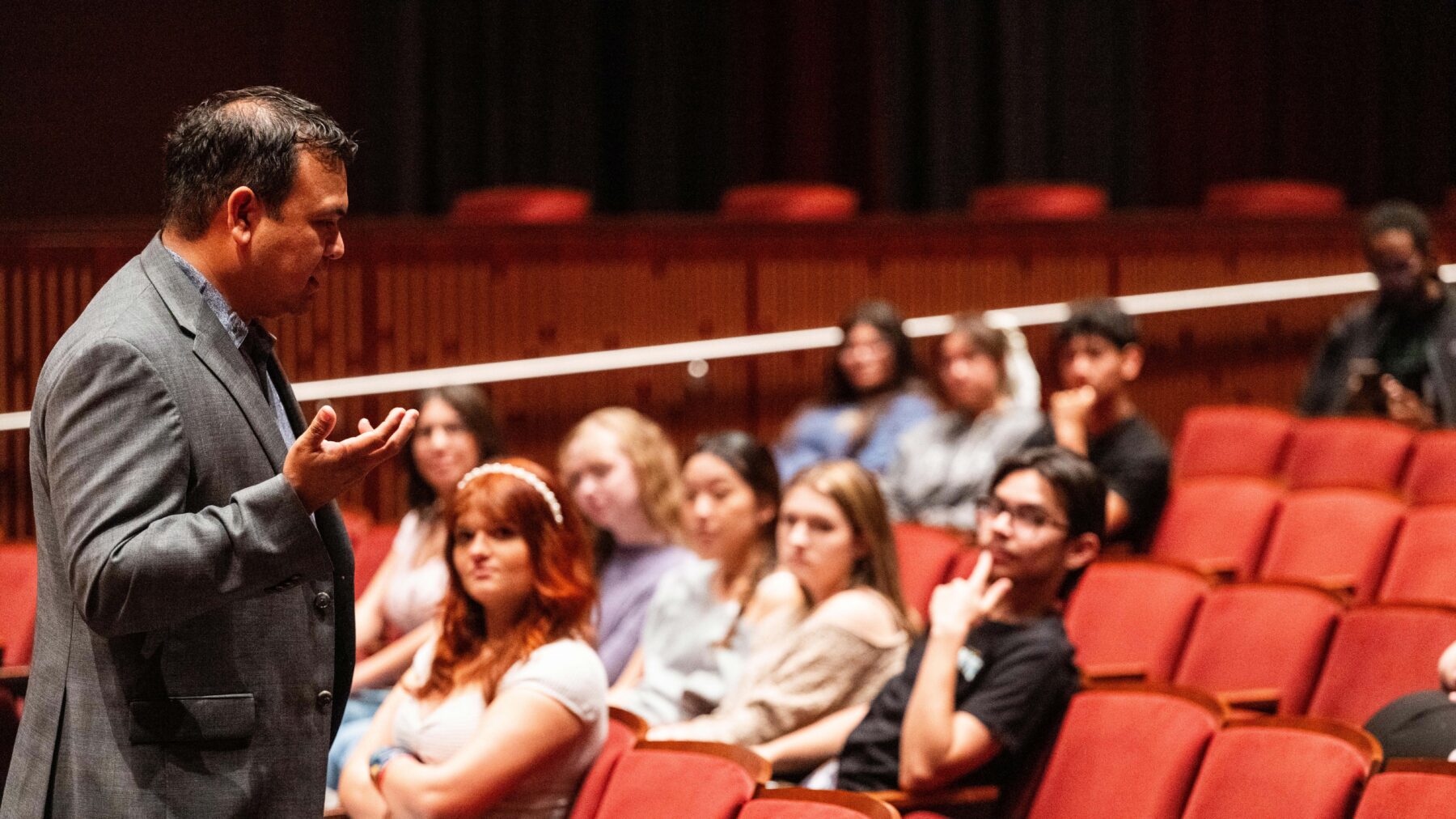 A visiting alum in a suit addresses students who are sitting in padded orange auditorium seats.