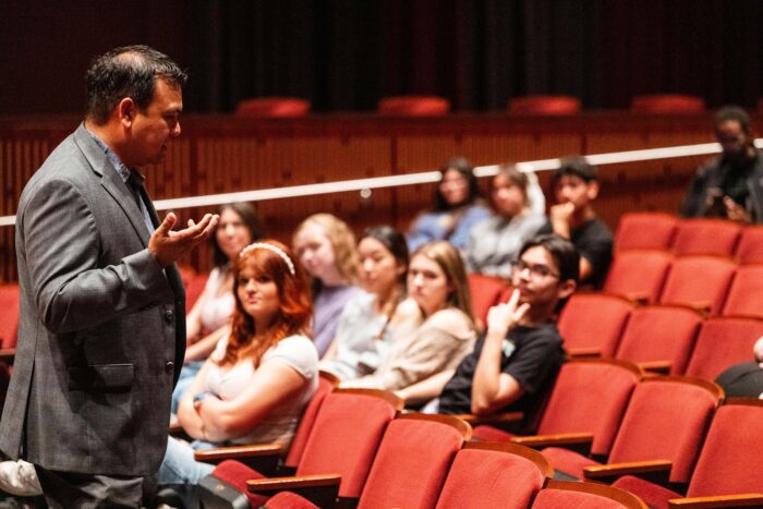 A visiting alum in a suit addresses students who are sitting in padded orange auditorium seats.