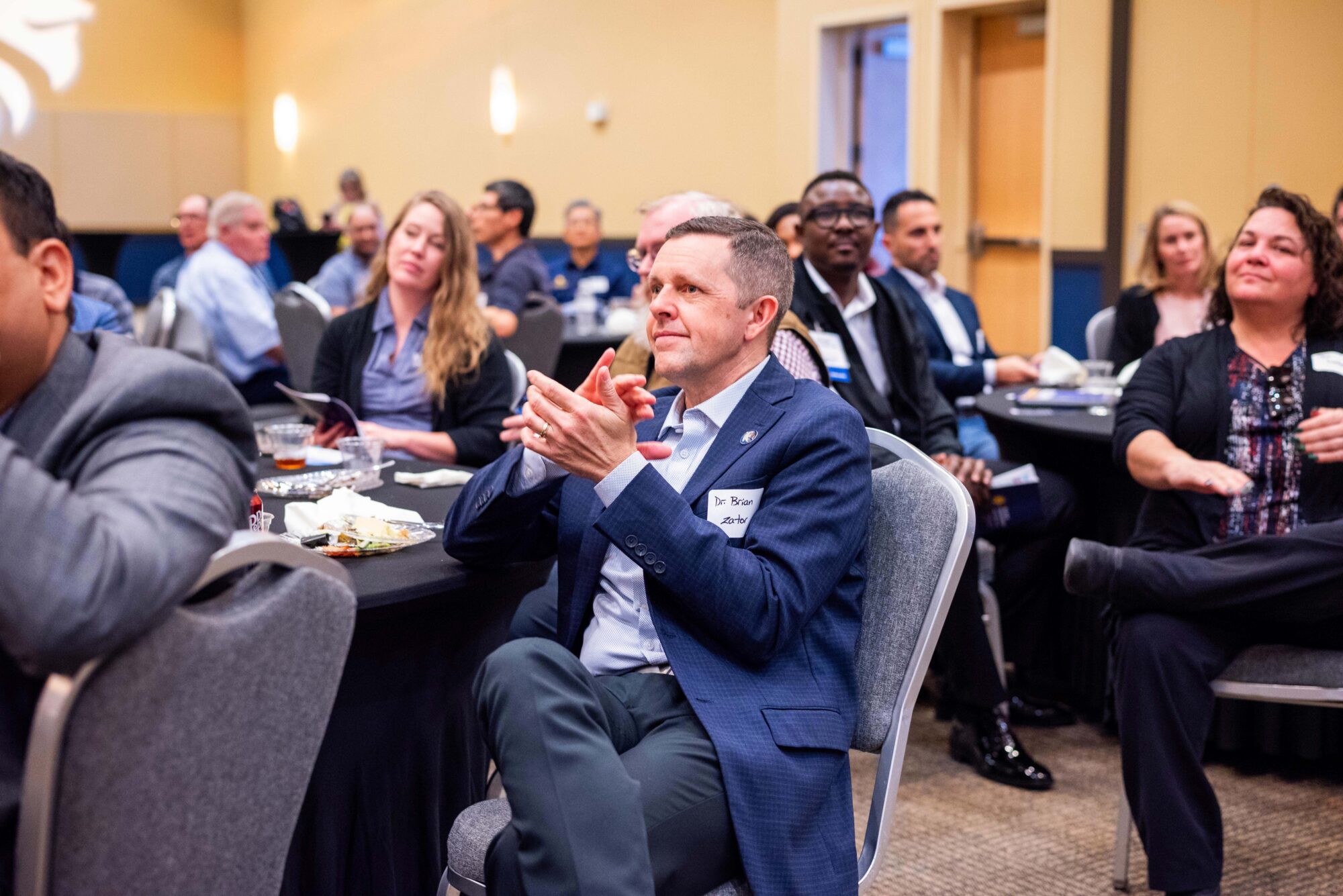 A crowd of people clap and look toward the front of the room. They are seated in padded chairs at tables with black tablecloths.