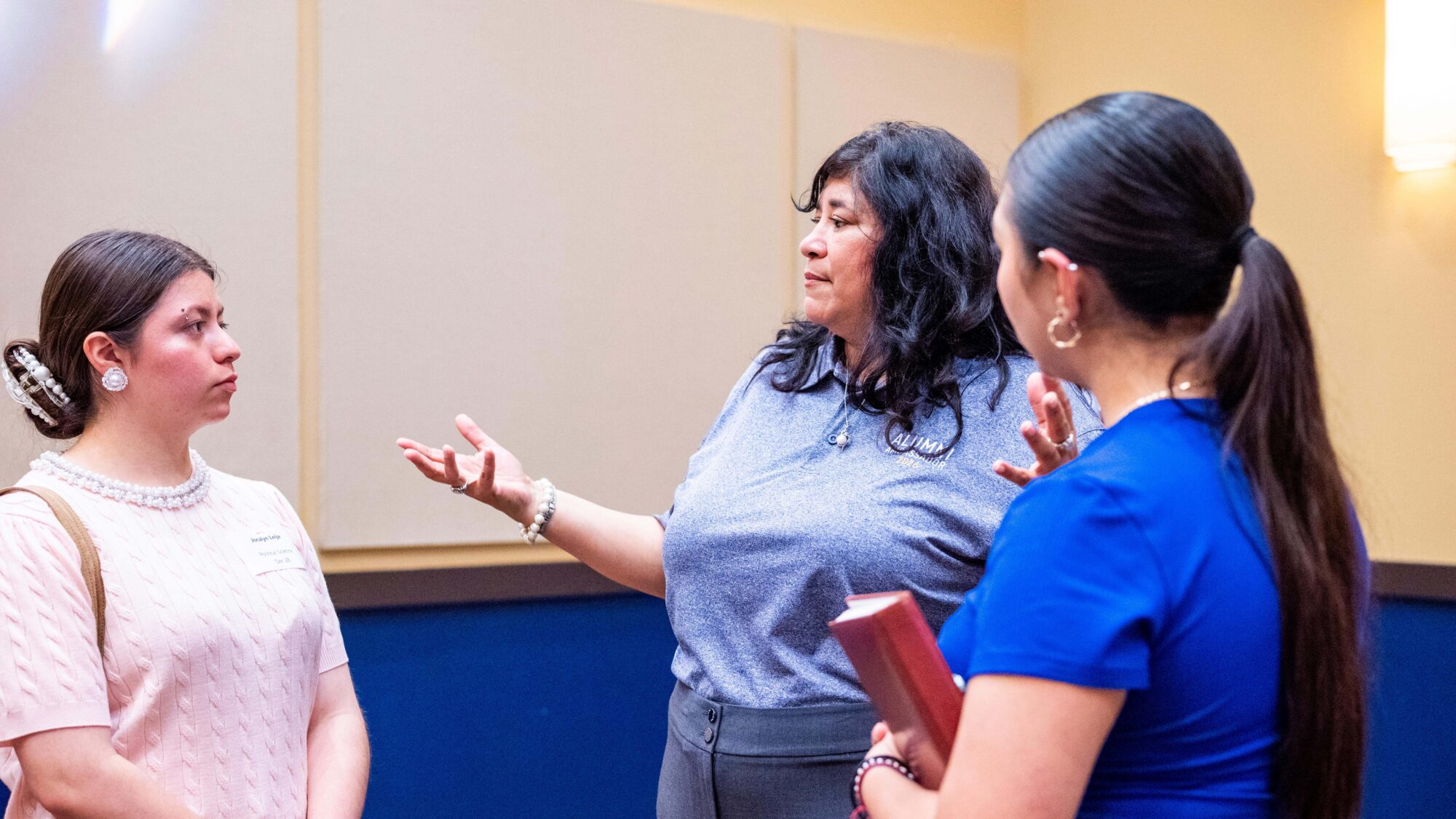 An alumnus stands between two students and speaks to them. Her right palm is upraised. The students listen to her speak with serious expressions on their faces.