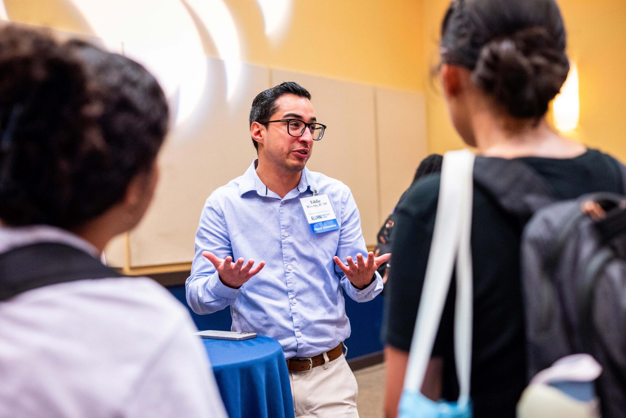 A visiting alum in a blue shirt and black glasses speaks to students. He is facing the camera and his palms are facing up in the air. He has a slight smile on his face. The students are facing away from teh camera and are listening to the alumnus as he speaks.