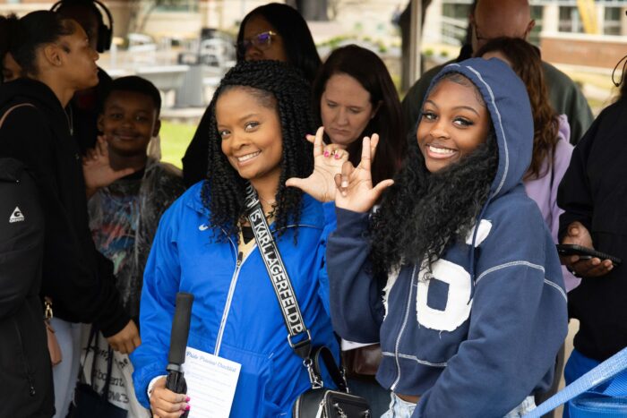 Two students in the foreground smile at the camera and make an "L" shape with their fingers. Several other students are shown in the background of the photo.