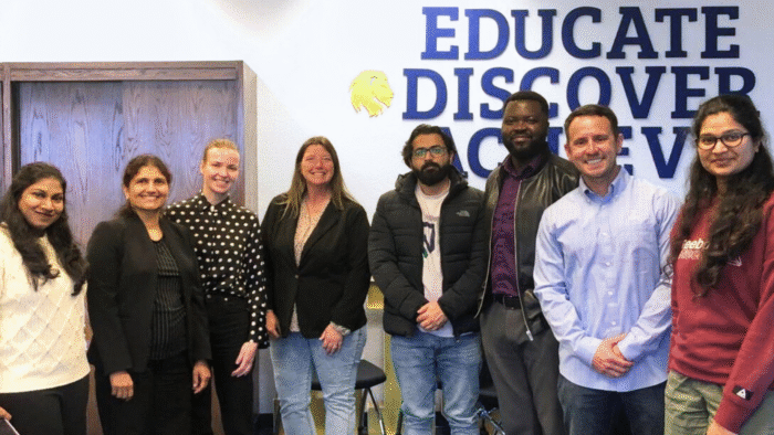 A group of people take a photo in front of a wall decorated with the slogan "educate, discover, achieve."