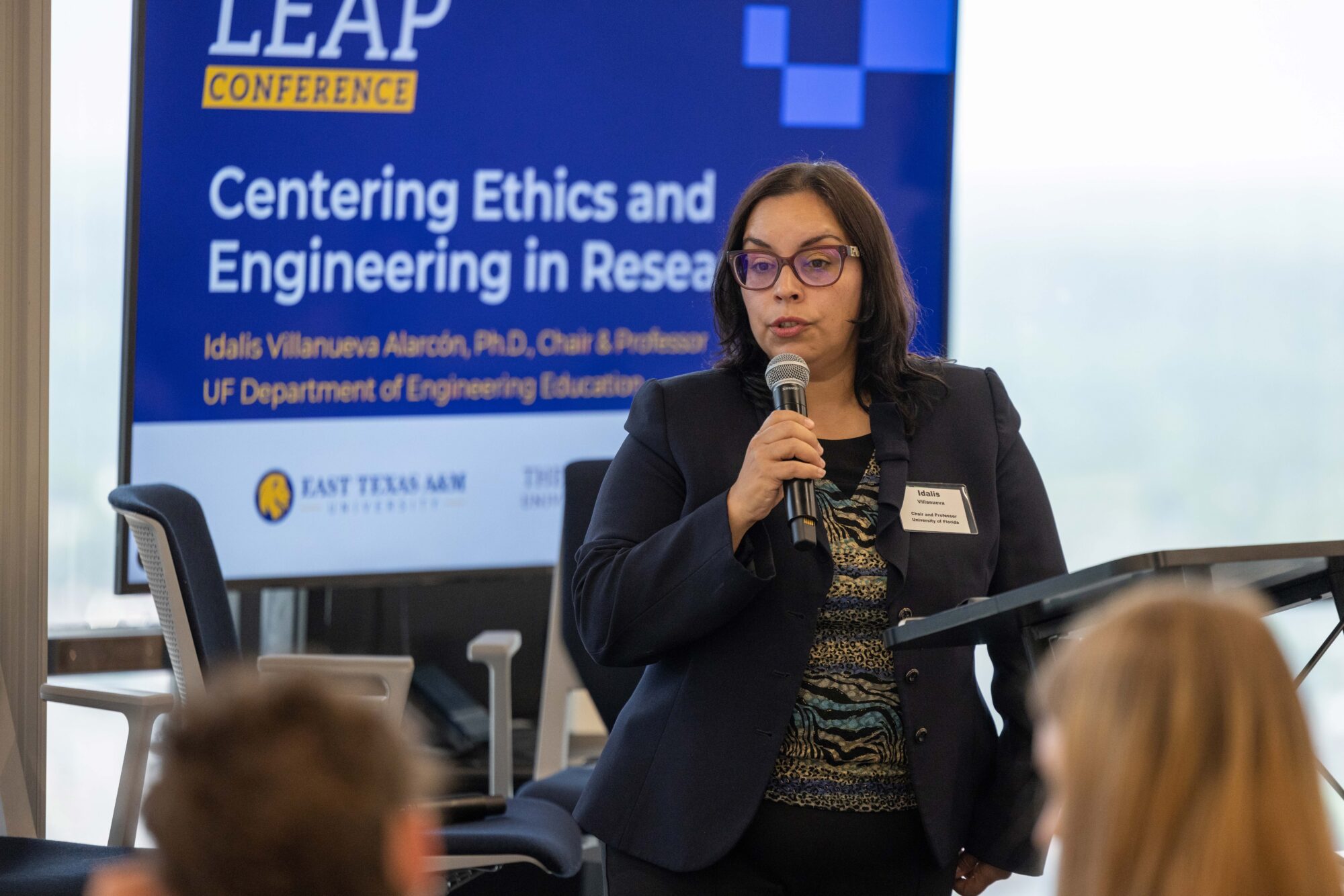 A woman in a black suit jacket  and red glasses holds a microphone and stands in front of a blue screen. She speaks to the crowd.
