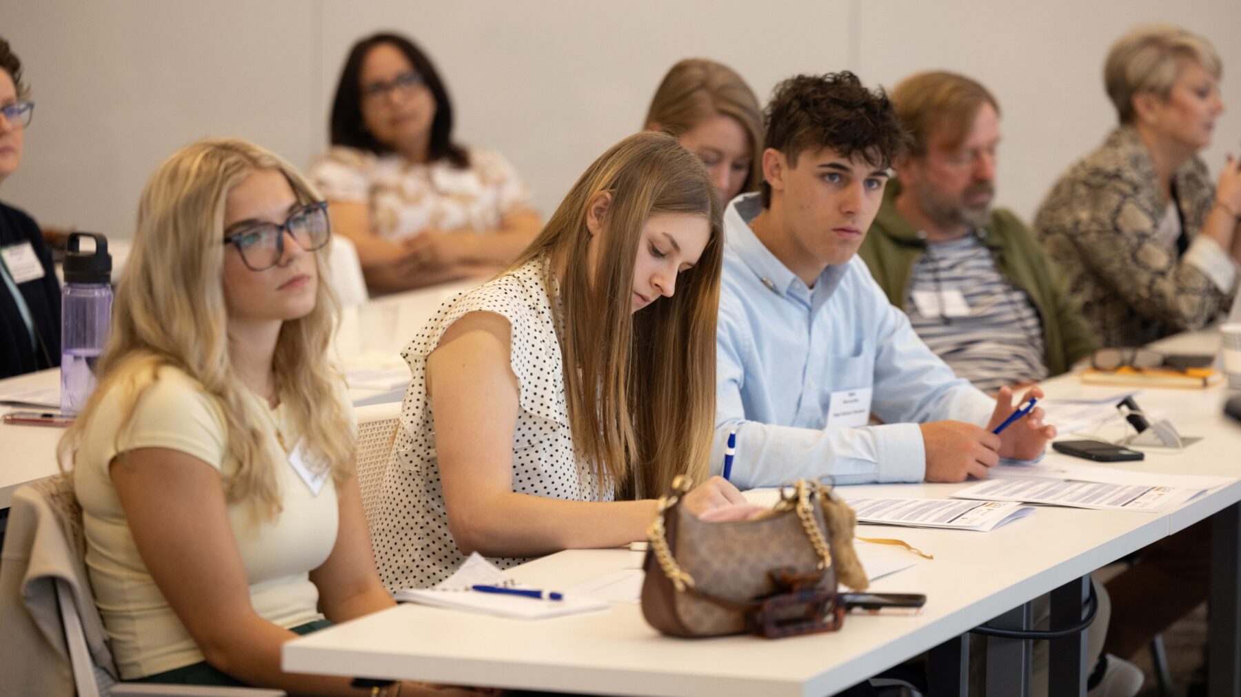 Around six people sit in two rows of tables with their faces forward, appearing to listen. One person has his elbows on the table.