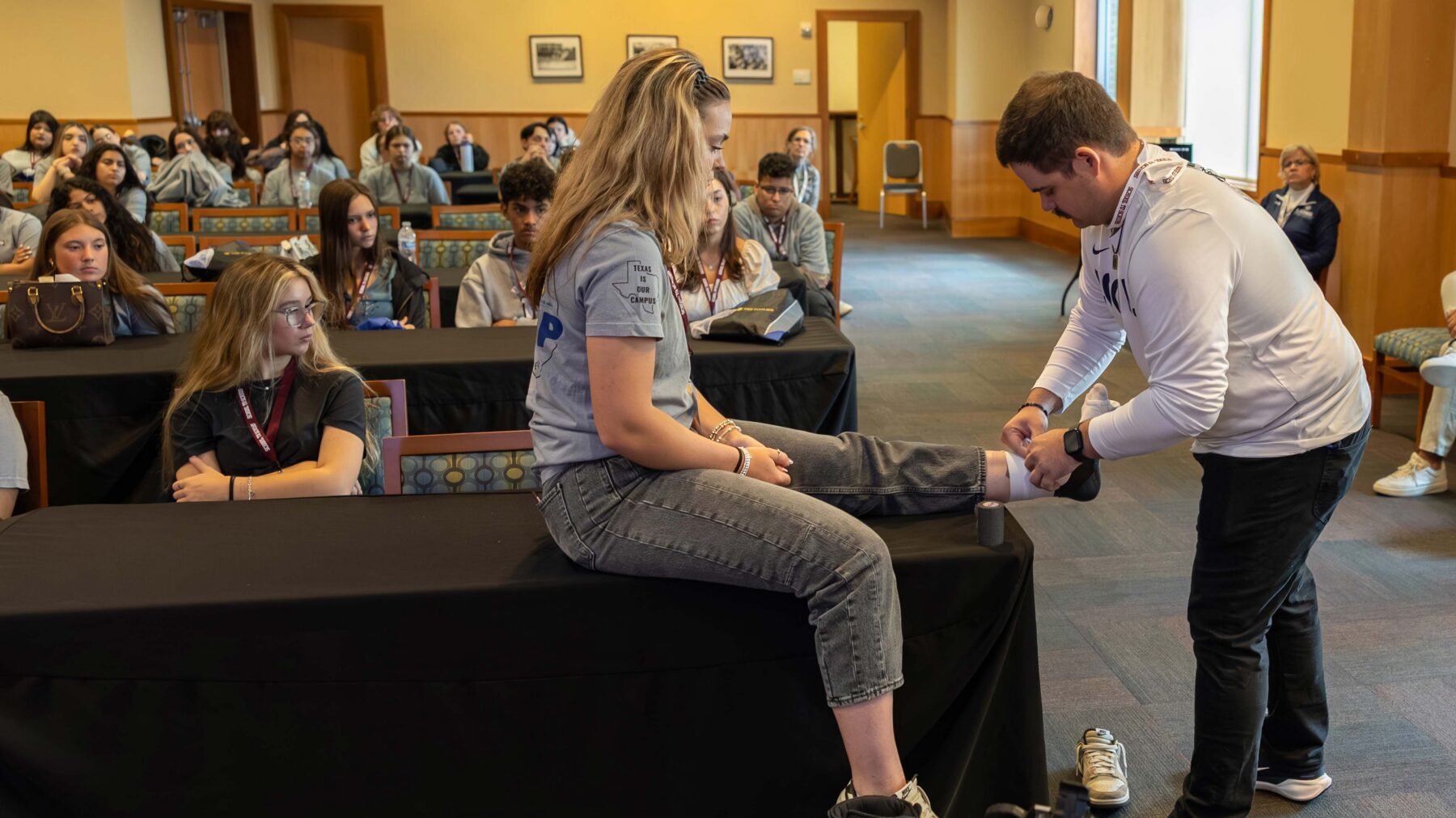 Classroom of students, with one girl sitting on a table having her ankle bandaged.
