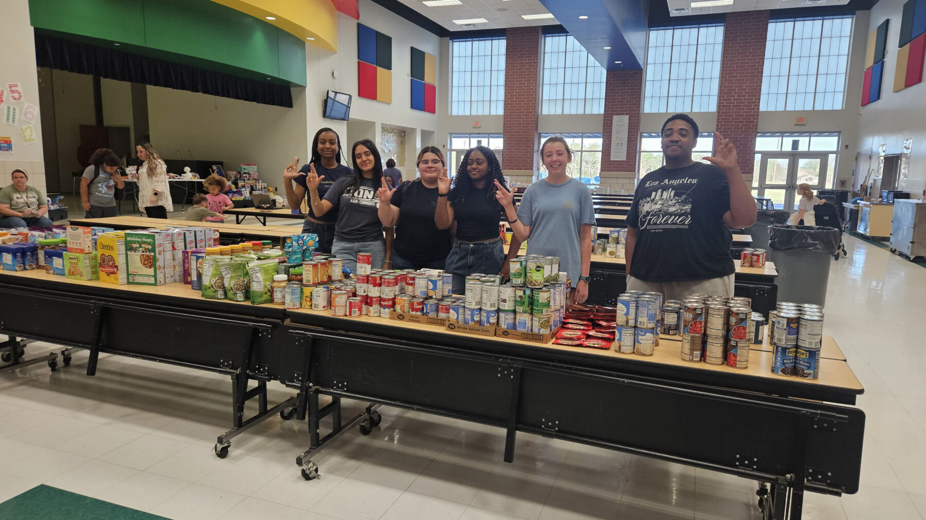 Several people stand behind cafeteria tables filled with canned goods and other groceries. The setting is a high school cafeteria.