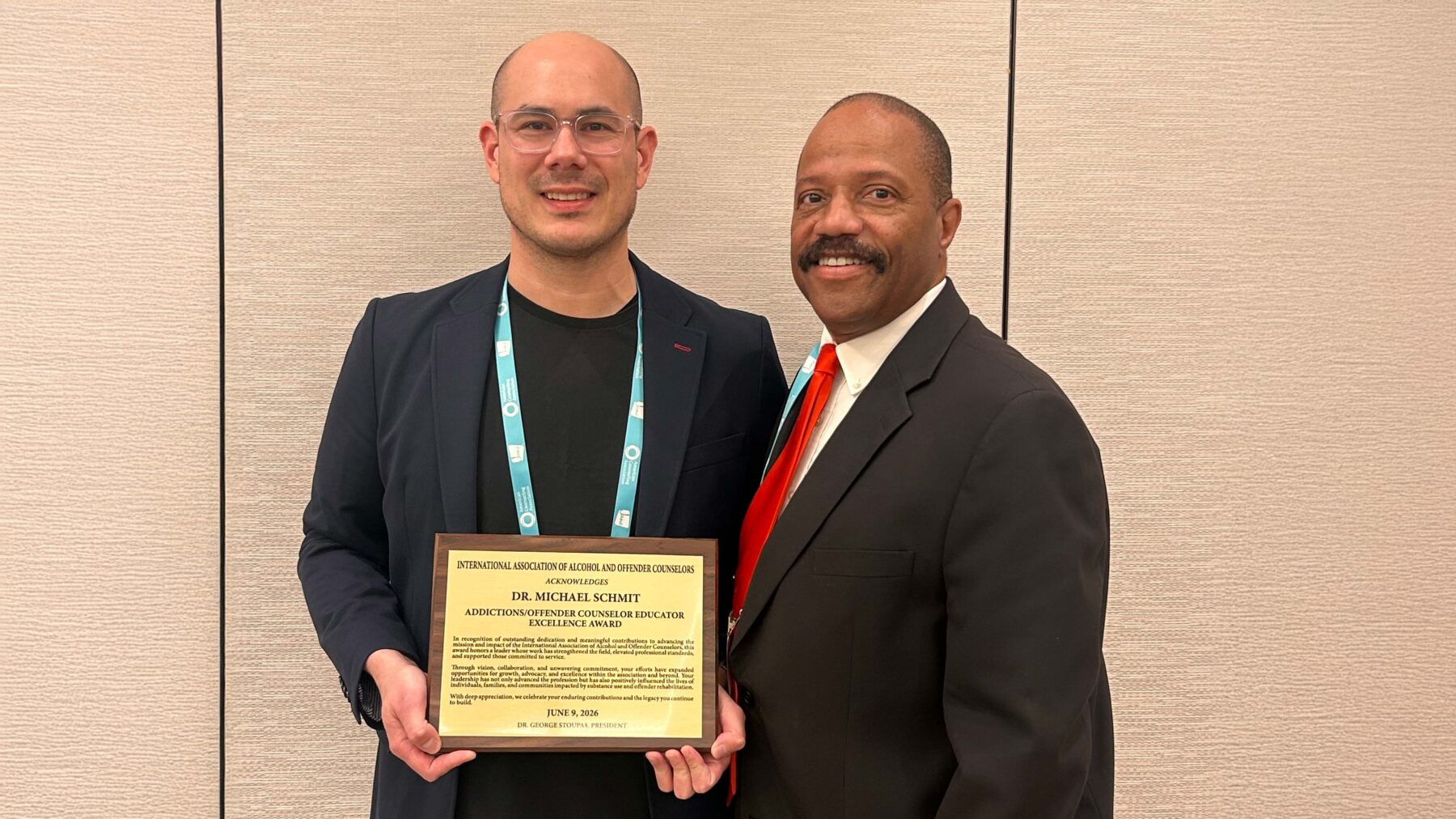 Two men stand against a wall with one of them holding a certificate of an award.