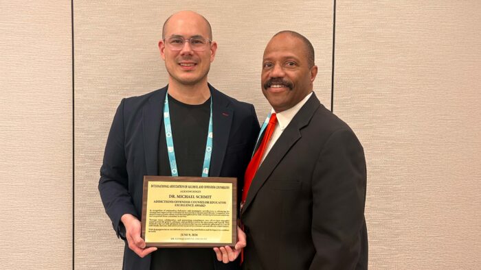 Two men stand against a wall with one of them holding a certificate of an award.