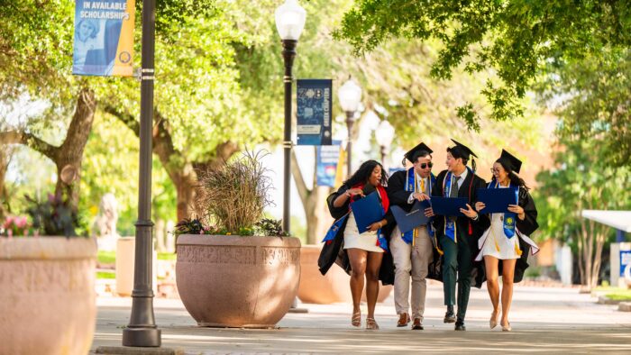 Photo for decorative purposes: Four students in black graduation gowns and black graduation caps walk together down the sidewalk, facing the cameral.