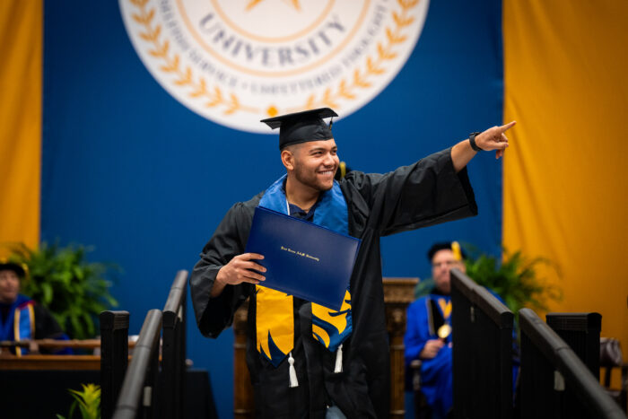 Male graduate on stage waving to family at graduation.
