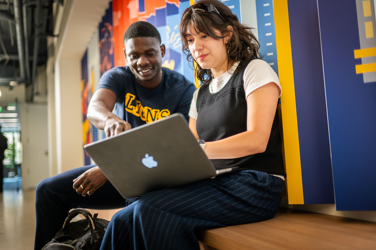 Two students sit on a bench inside the university looking at a shared laptop together.