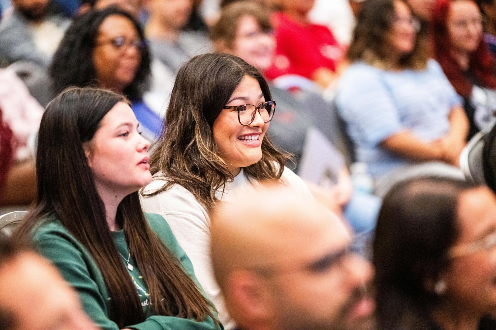 Two students in a crowd (which is out of focus), look forward toward the front of the room. One student is smiling.