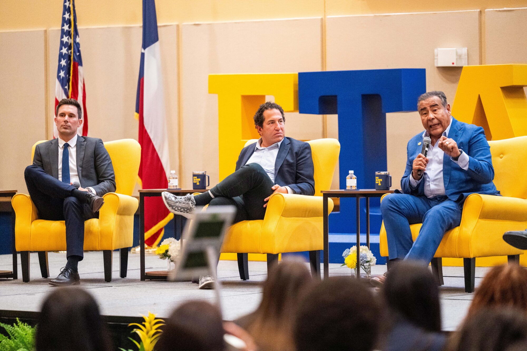 Three people sit on yellow armchairs. They are wearing suits. The person on the right is speaking into a microphone and the other two are listening. They are sitting up on a stage.