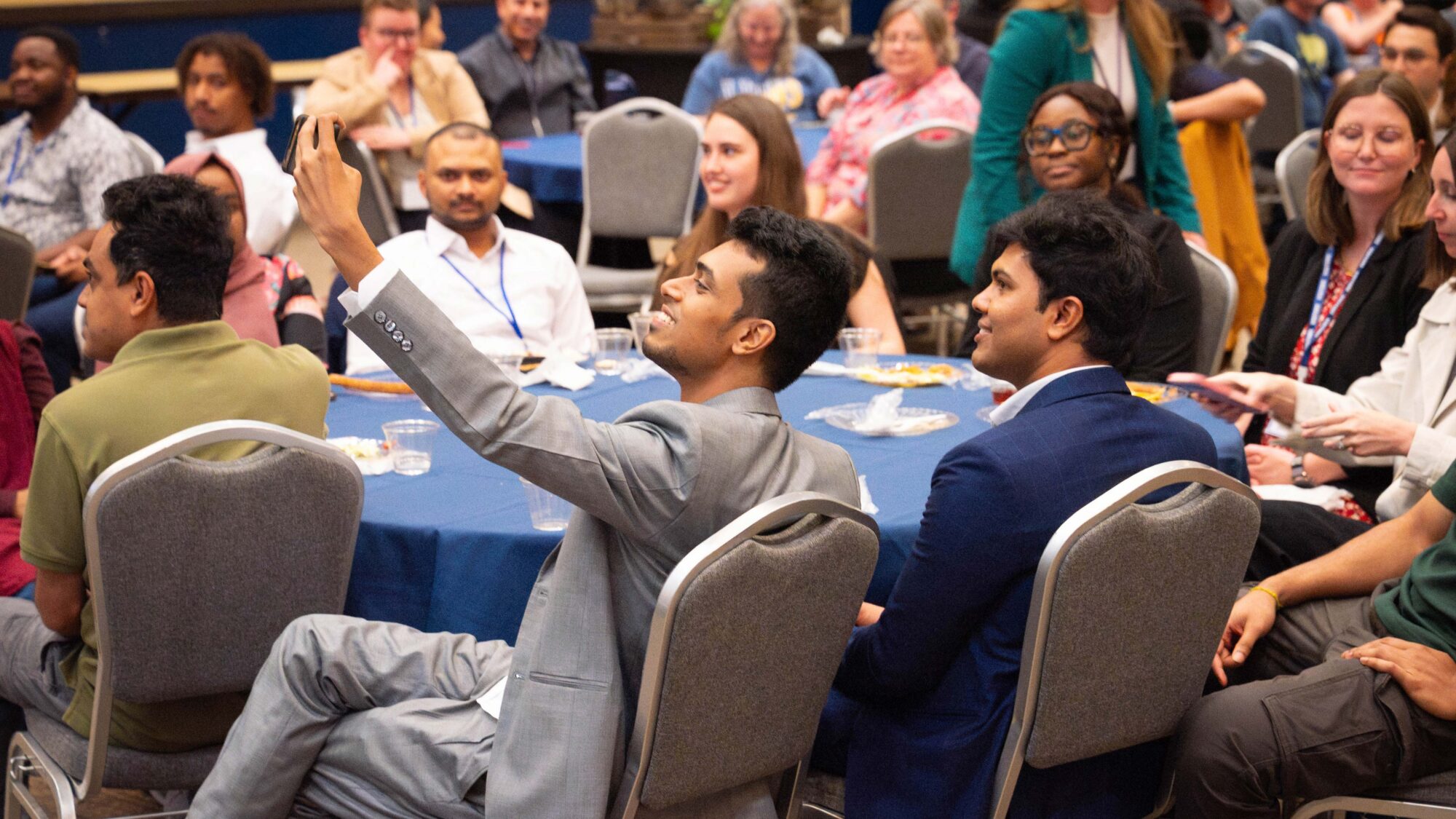 A group of around nine people are sitting around a table with a blue cloth over it. The person closest to the camera has his arm lifted to take a selfie with the person in the chair next to him. 