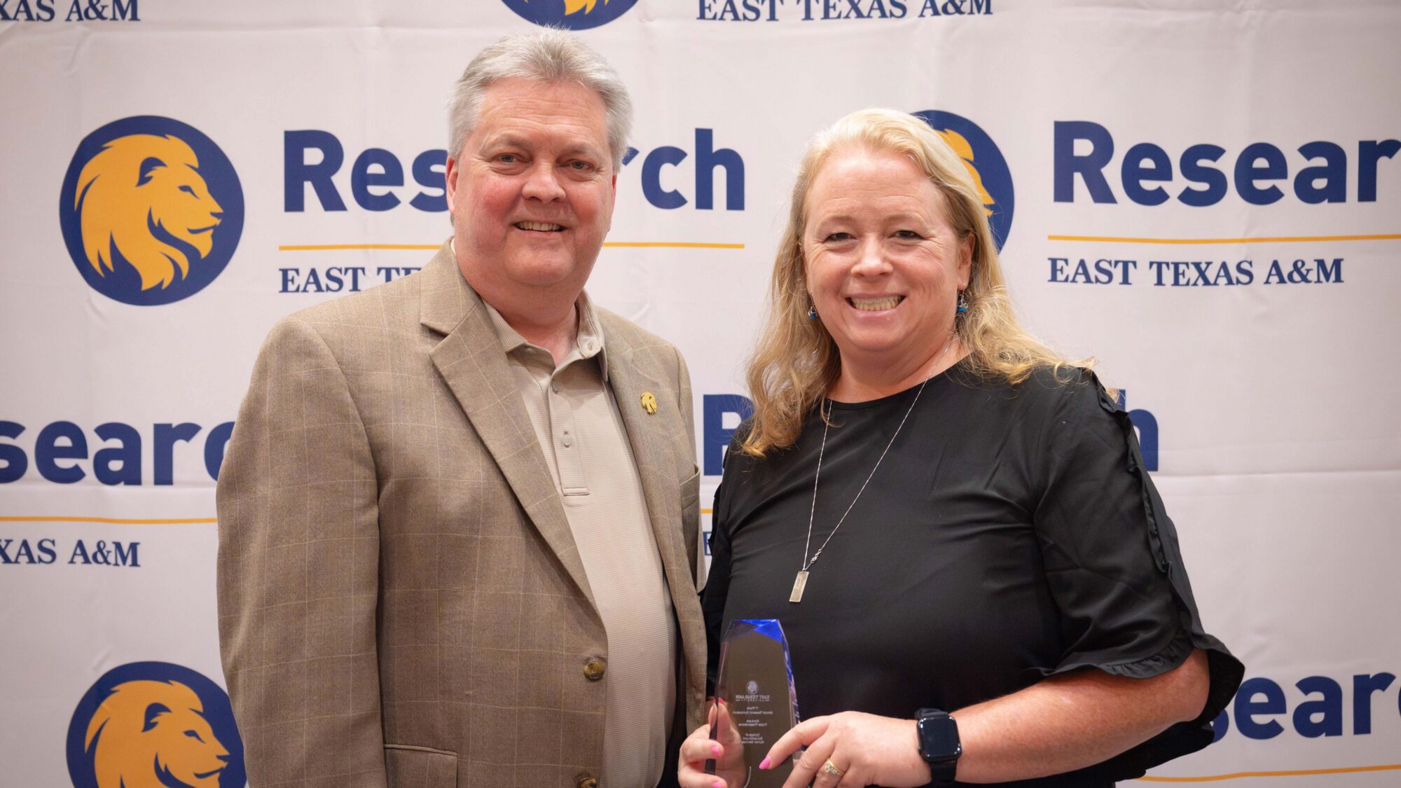 Two people smile for the camera. The woman on the right is holding an award. The backdrop says "Research" and includes a lion head logo.