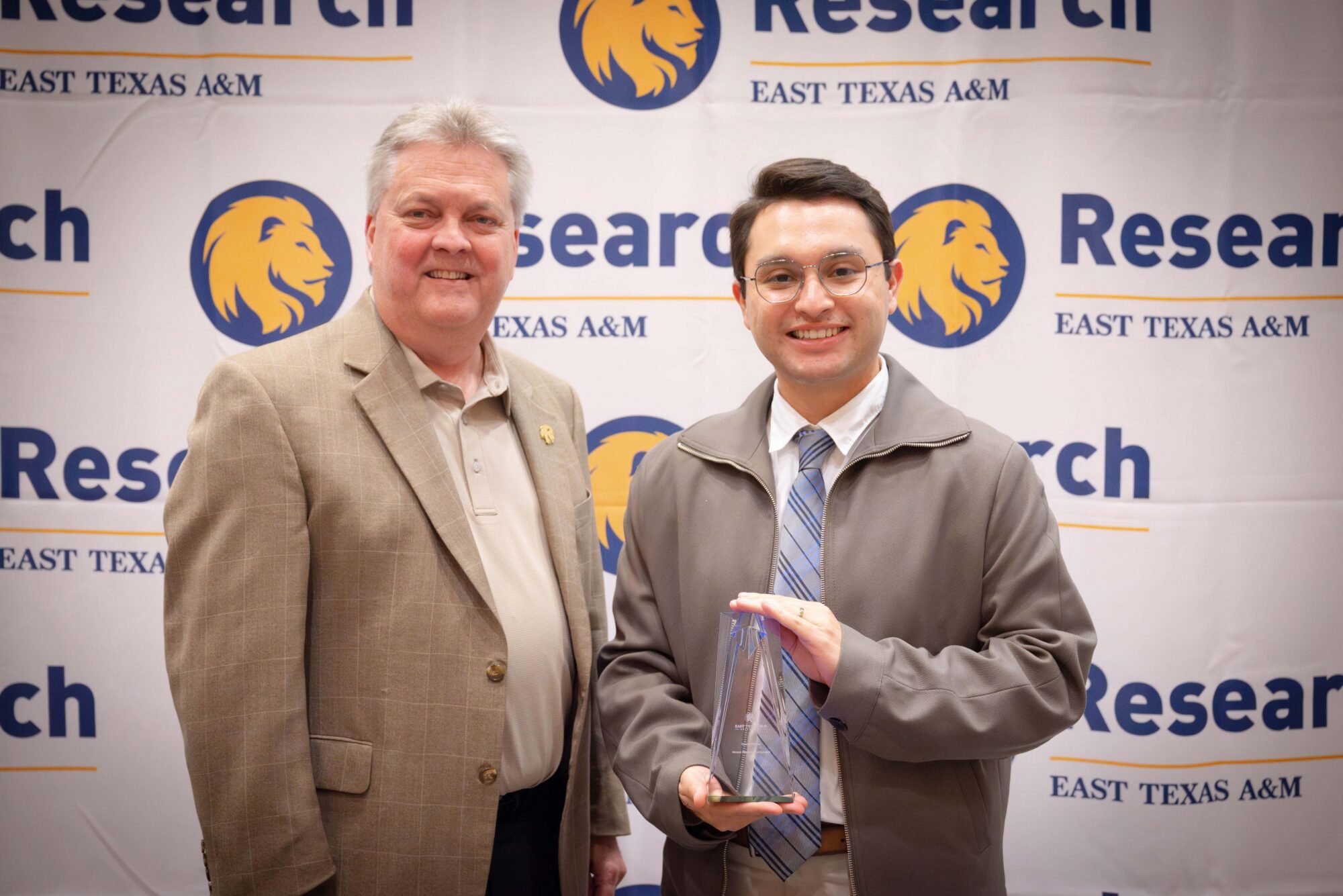 Two people smile for the camera. The man on the right is holding an award. The backdrop says "Research" and includes a lion head logo.