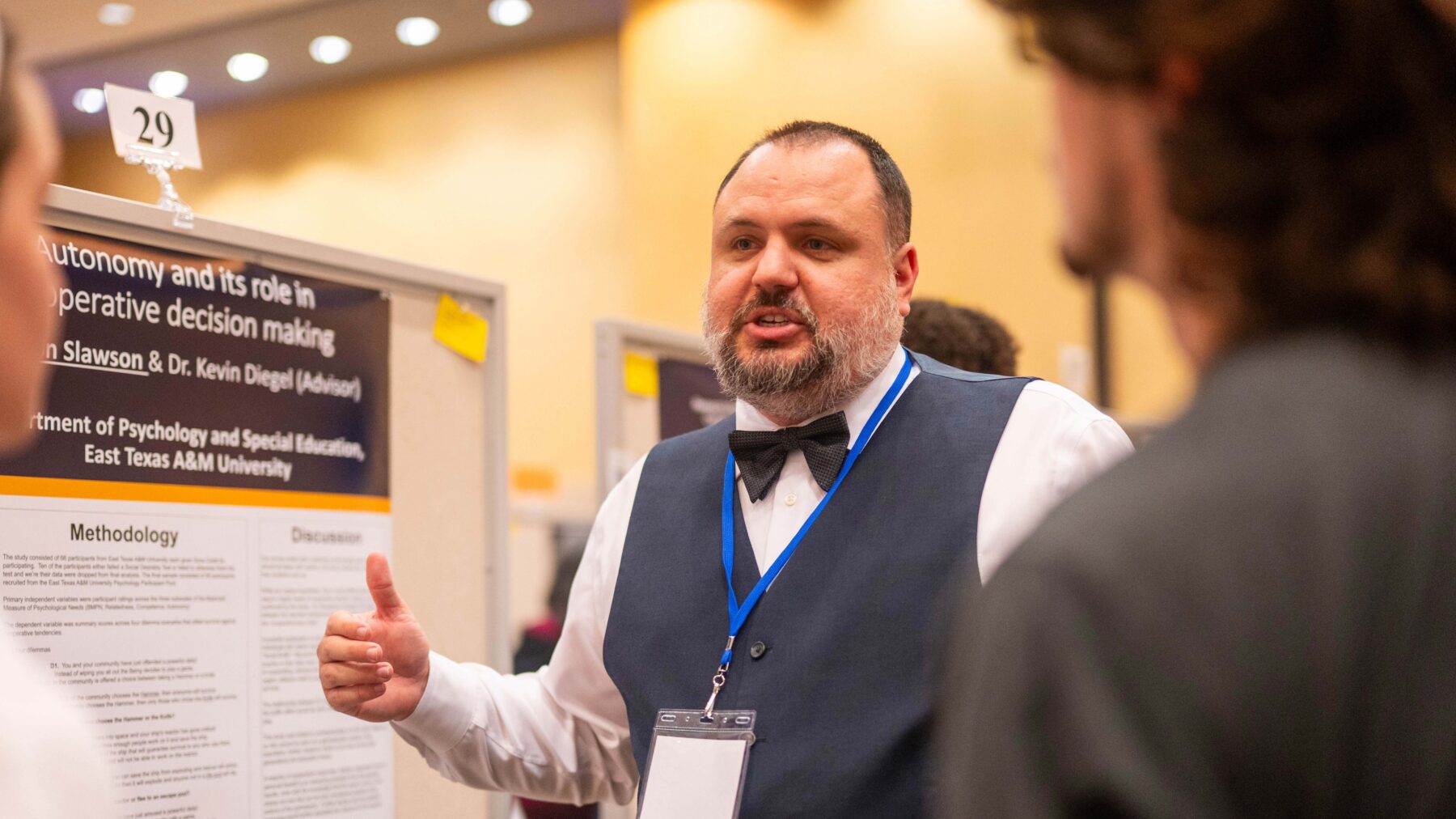 A student researcher speaks to two people as he stands in front of a display board with information printed about his research.