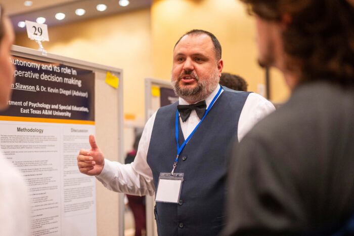 A student researcher speaks to two people as he stands in front of a display board with information printed about his research.