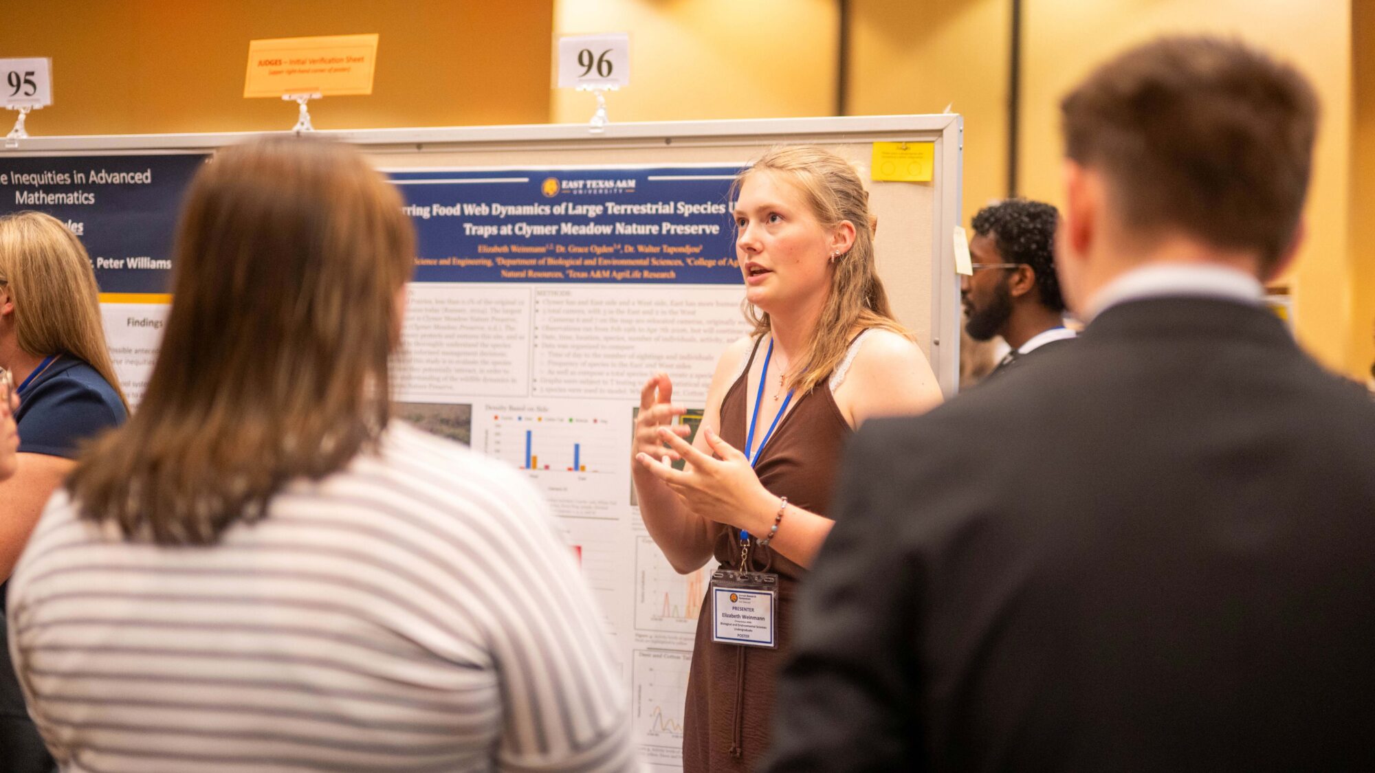 A student stands in front of her display board and speaks. Her palms are pointed upward, and she is looking up. Two people, with their backs to the camera, listen to her presentation.