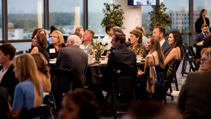 Several people sit around a round table, covered in a black cloth. They are dressed in formalwear. The Dallas skyline appears behind them.