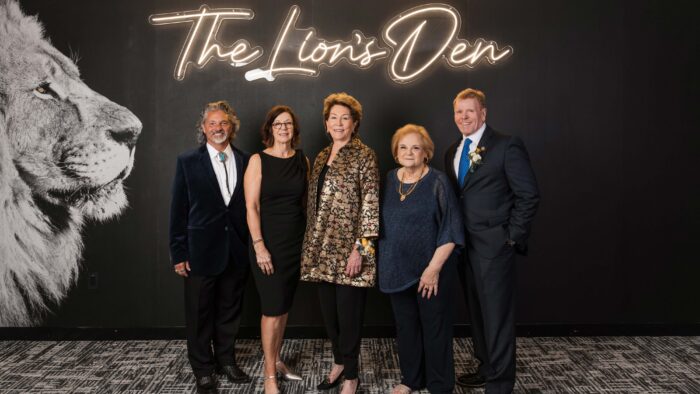 Five award recipients stand in a row with a black background and a lion head on the left side and the words "The Lion's Den" in lighted gold letters above them.