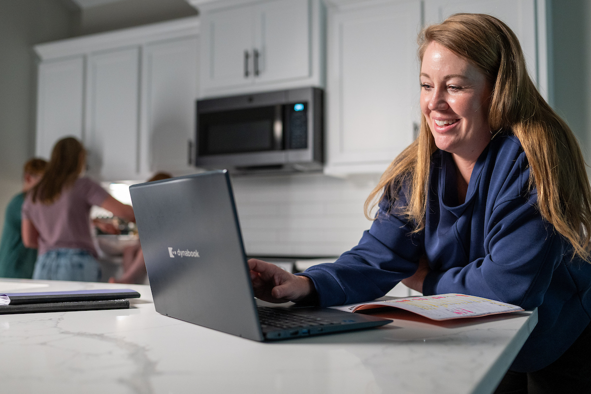 Woman uses her laptop at a kitchen counter with her children in the background.