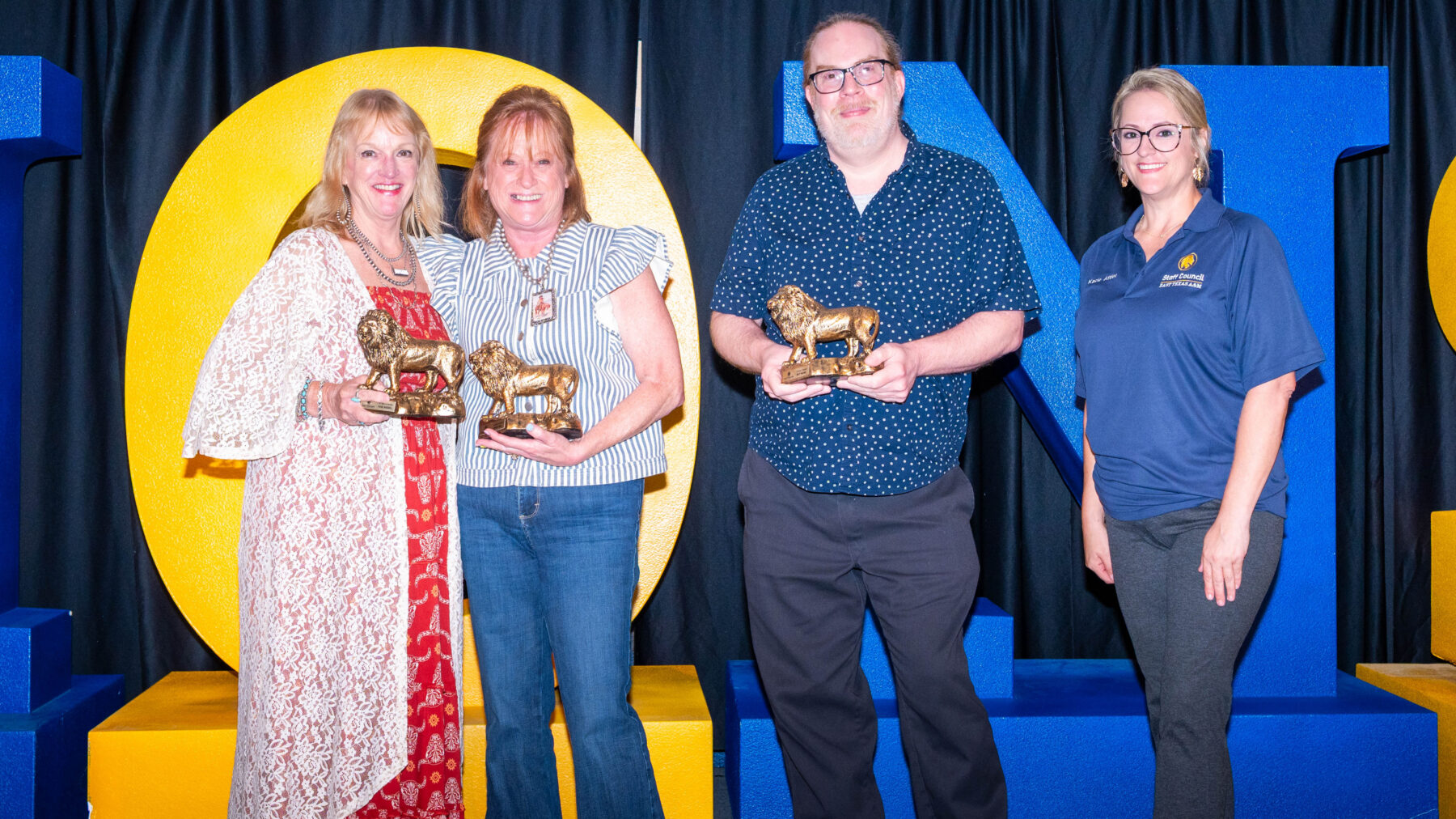 Matt Meinke, second from the right, stands beside two award recipients and another staff member wearing a blue polo shirt. Four people stand in front of large blue and yellow letters spelling “LIONS.” Three of them hold gold lion‑shaped awards.