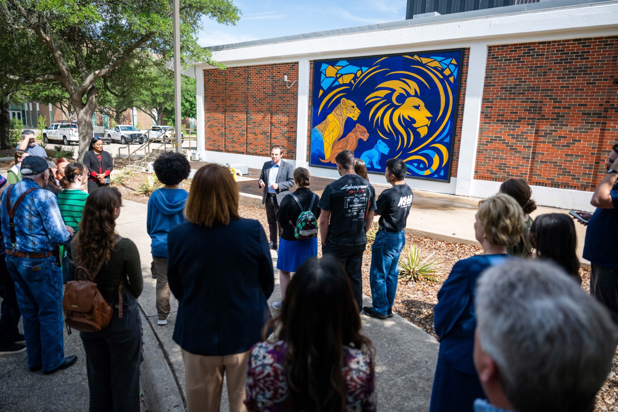 Onlookers look toward the speaker, in a suit, at the front of the group. Behind him is the mural mounted on a brick building featuring four Lions.