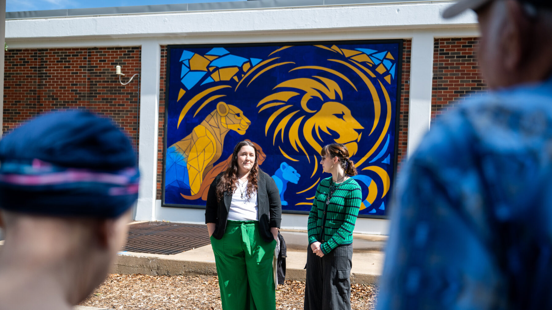 The two artists stand in front of their mural featuring four Lions. The artist on the left is wearing green pants and the artist on the right is wearing a green sweater with navy stripes. They are looking at each other and talking.