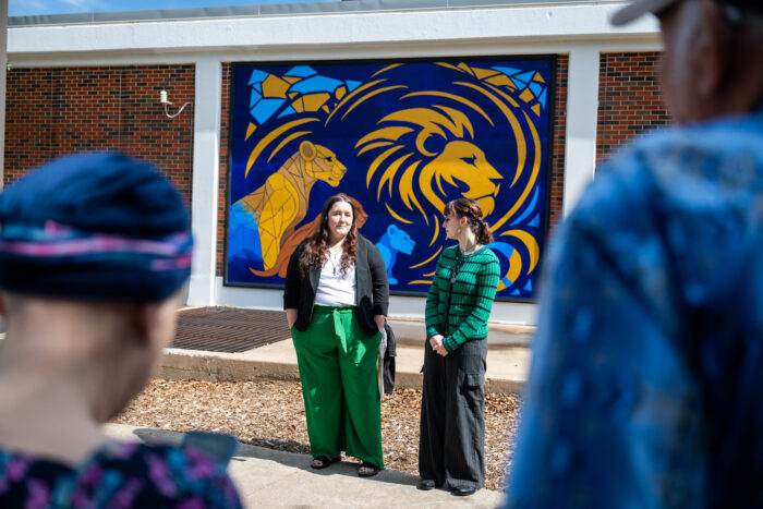 The two artists stand in front of their mural featuring four Lions. The artist on the left is wearing green pants and the artist on the right is wearing a green sweater with navy stripes. They are looking at each other and talking.