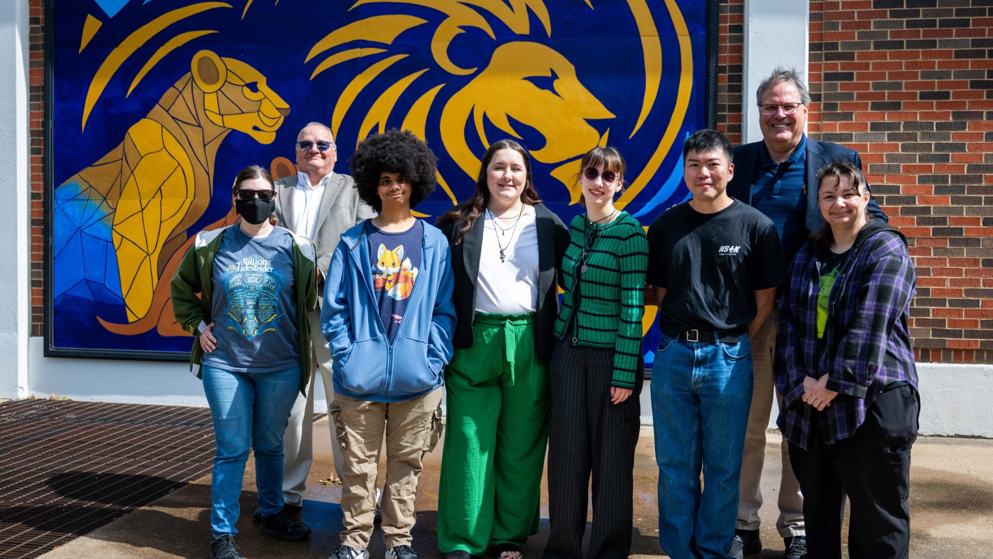 A group of eight people stand in front of the Lion mural, mounted on a brick building. The group smiles at the camera.