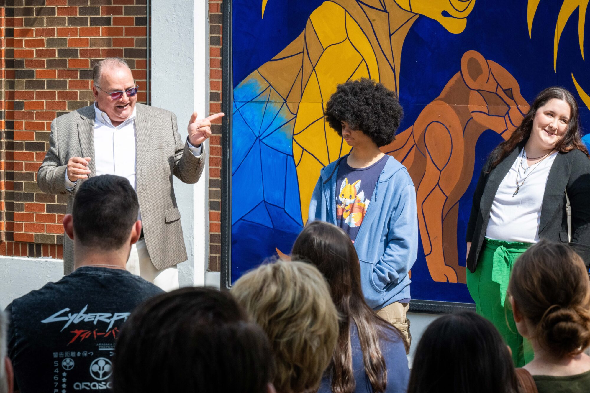 Three people stand in front of the mural. The man on the left is speaking and making a pointing gesture with his finger. Six people from the audience are also shown from the back, listening to the speaker.