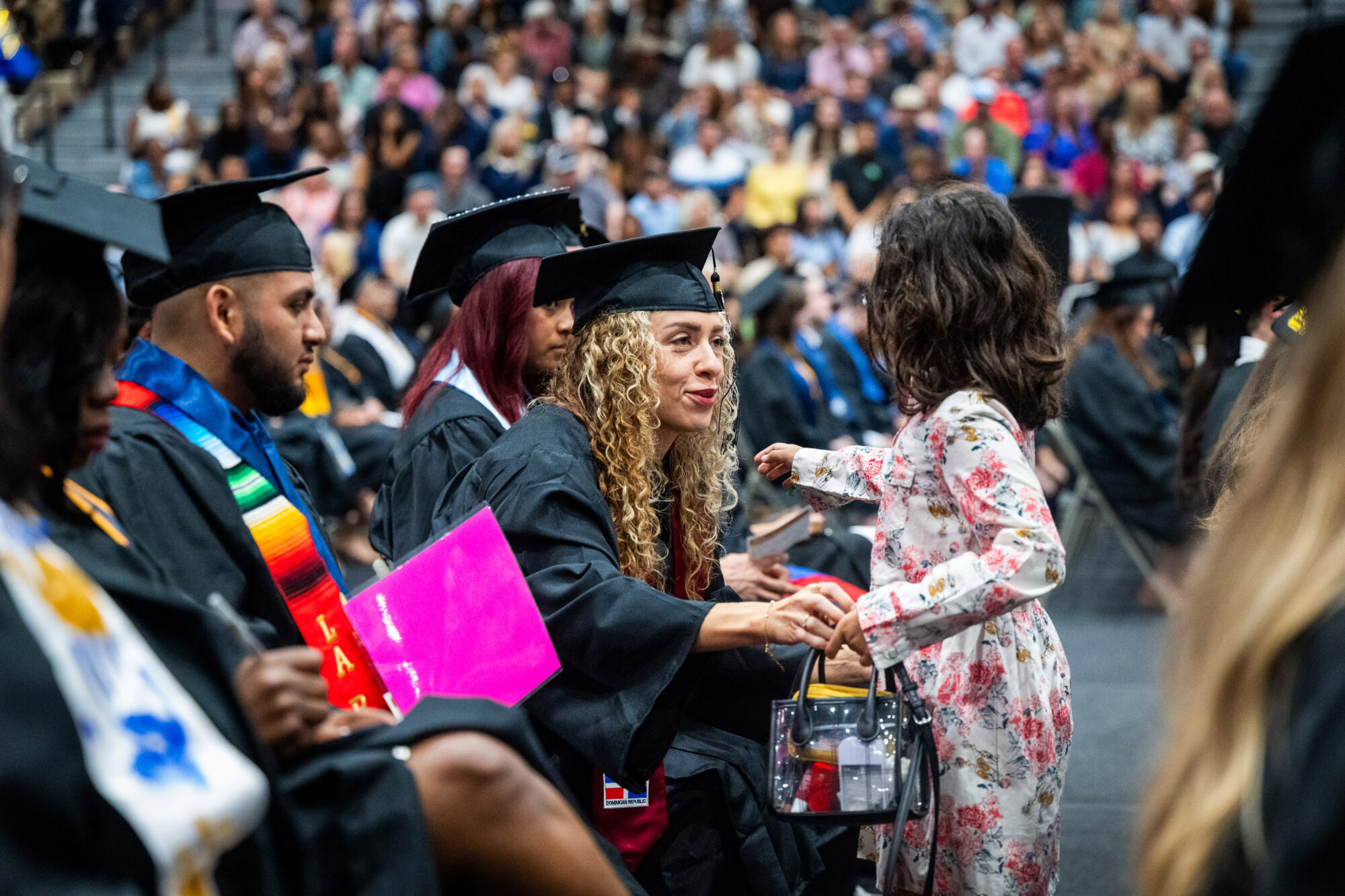 A row of graduates sit in the auditorium. One of the graduates is leaning forward with her arm outstretched to hug a young girl who is standing in front of her.