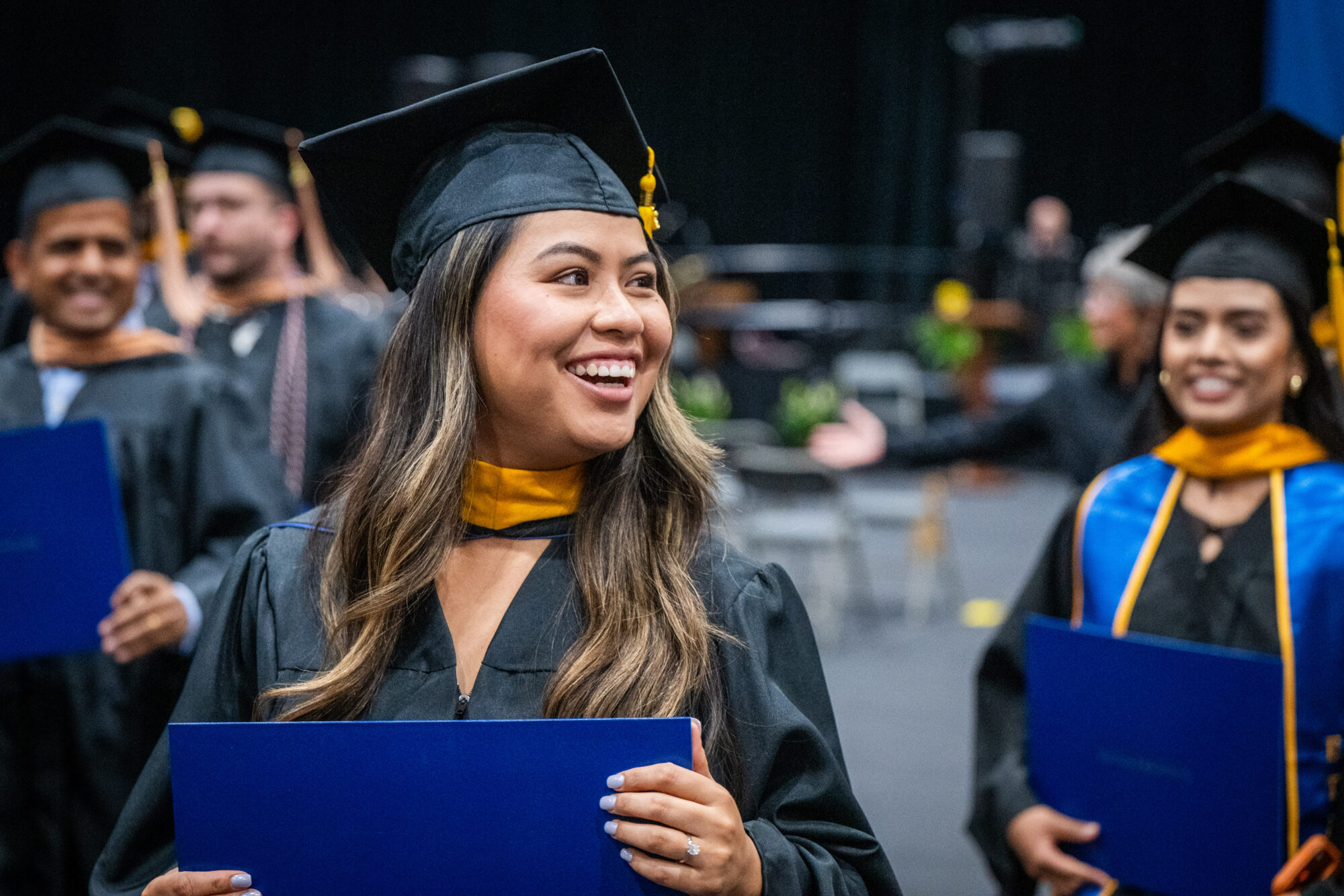A graduate in a black cap and gown turns her head to the side and smiles widely. She is holding a blue square document, her diploma. In the background, two other graduates look at her and smile.